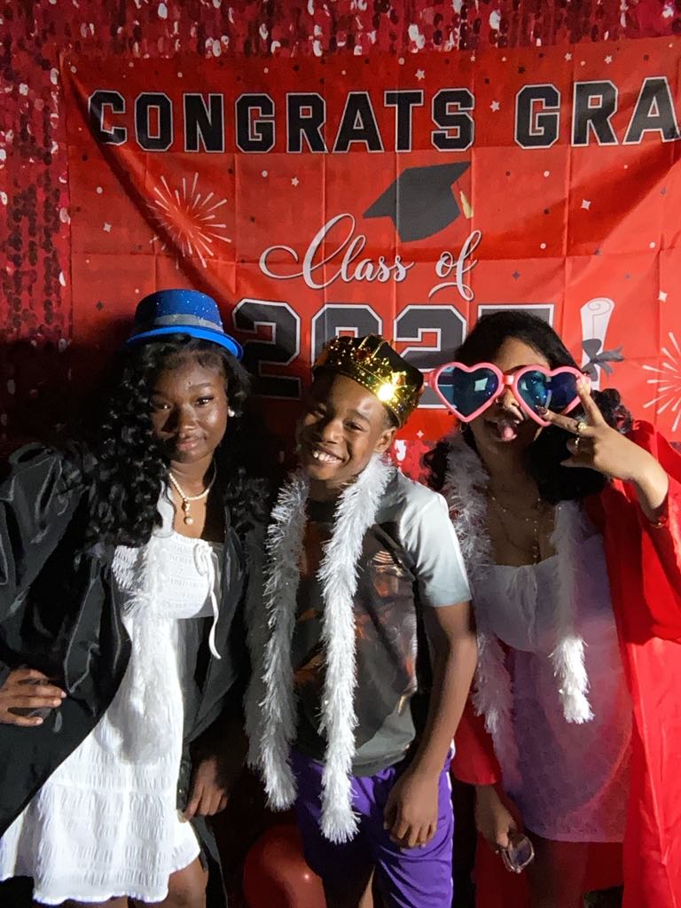 Three young people posing in front of graduation banner with festive accessories